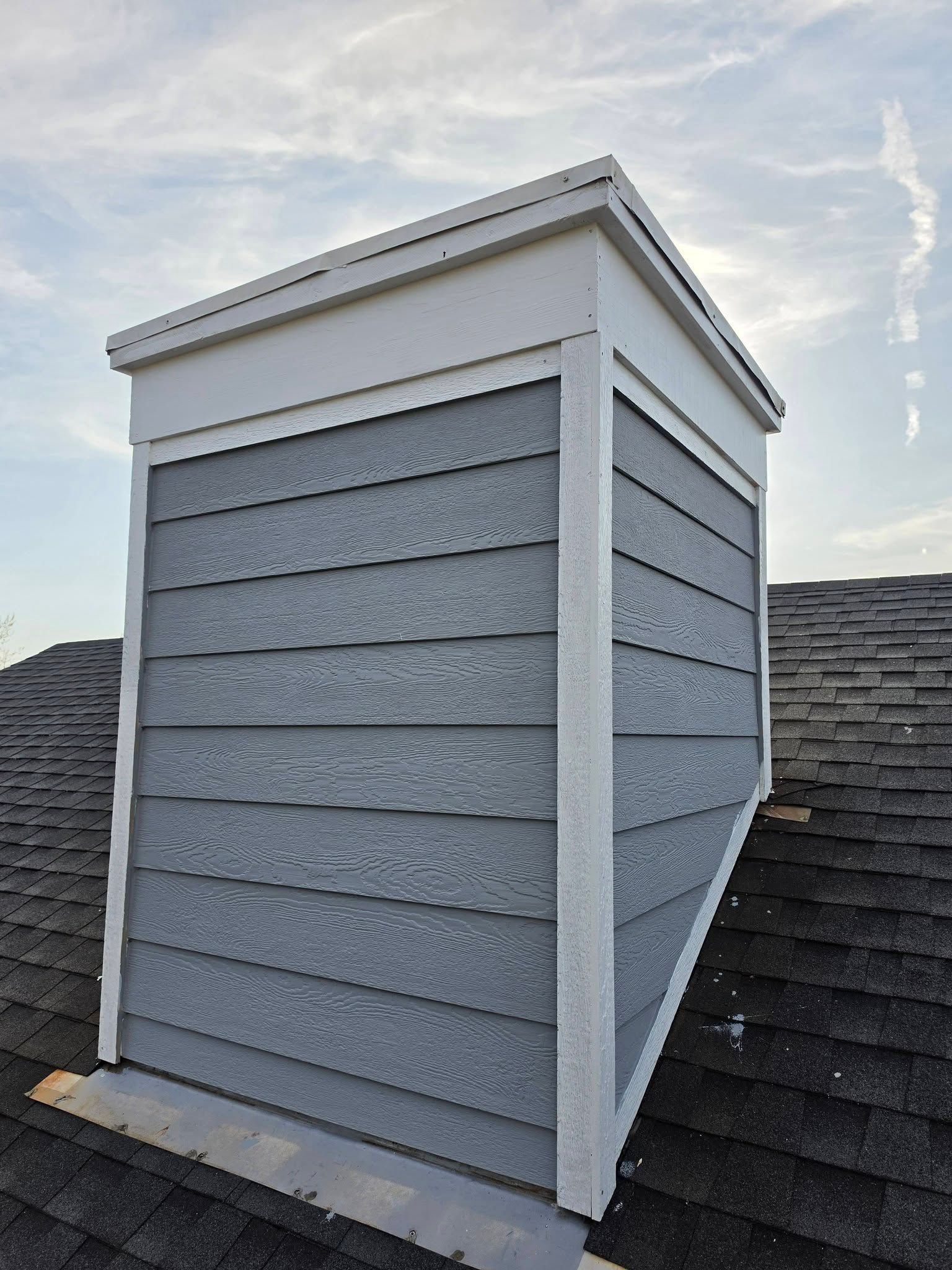 Gray dormer with white trim on a shingled roof against a cloudy sky