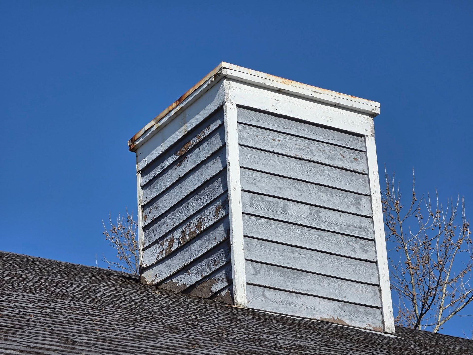 Gray rooftop chimney with white trim against a clear blue sky.