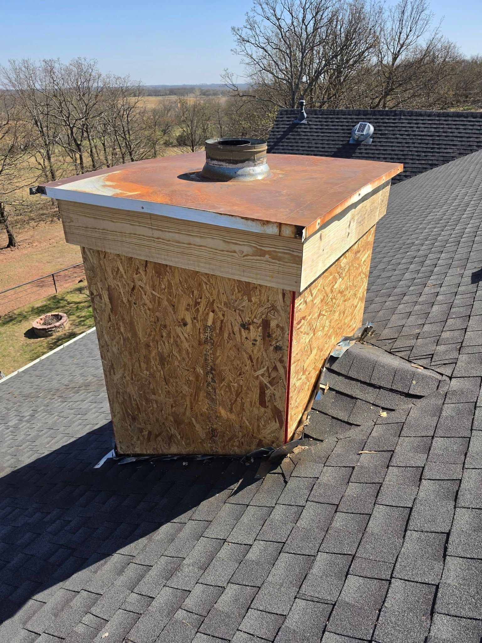 Rooftop chimney chase with rusted metal cap and shingle exterior on a sloped roof, against a rural backdrop