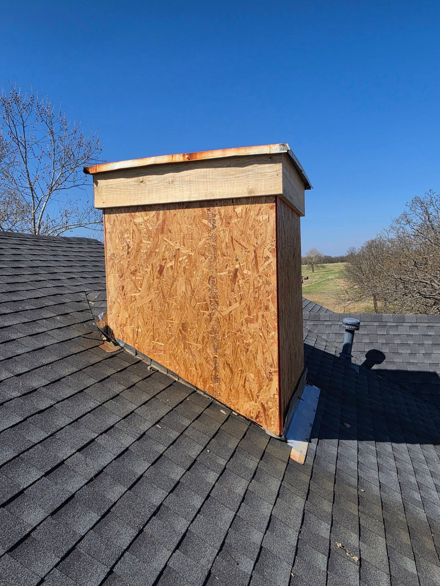 Rooftop chimney stack wrapped in plywood, set against a clear blue sky over gray shingles