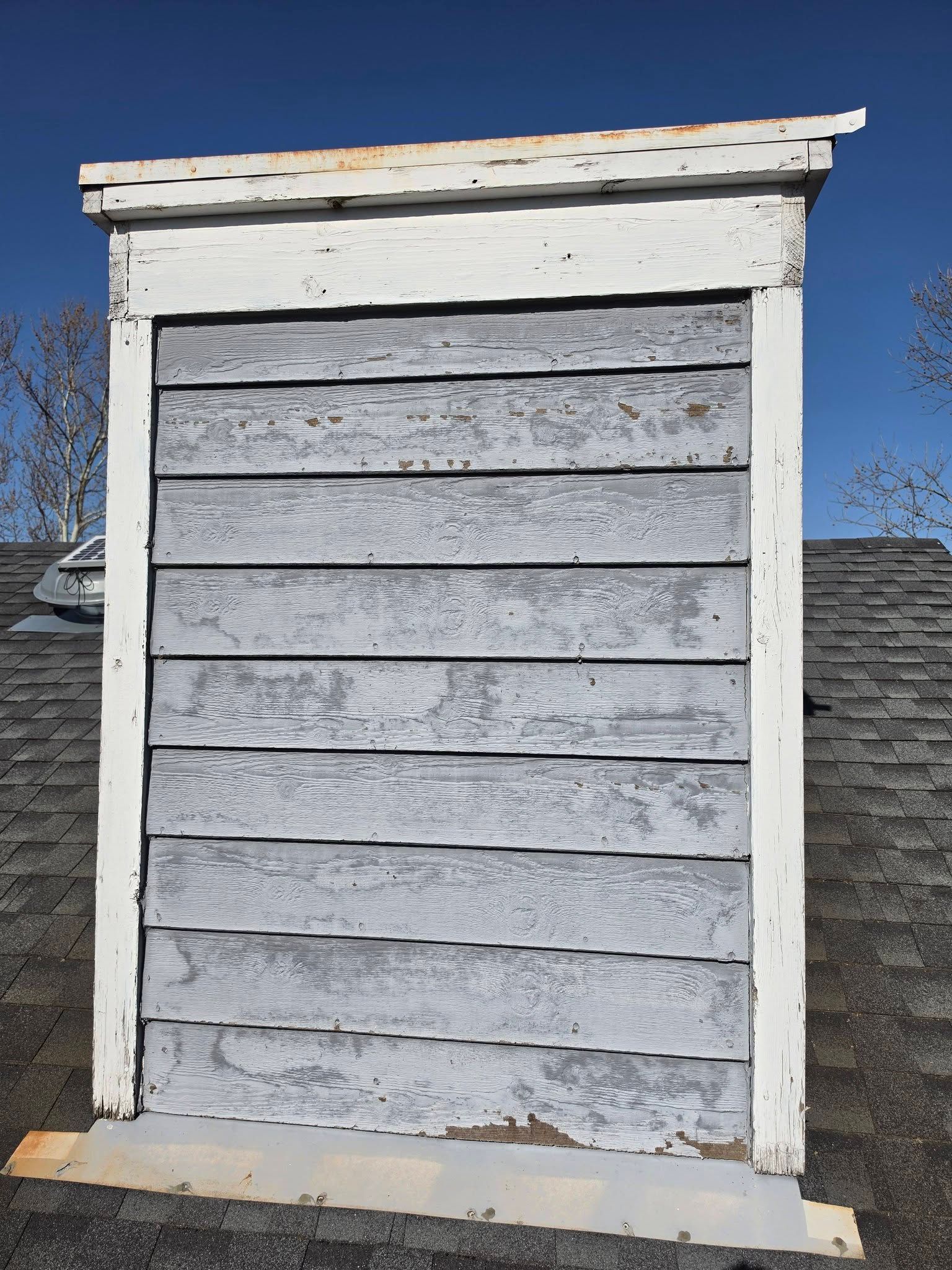 Weathered white wooden vent or small shed wall against a blue sky, with peeling paint and gray siding.