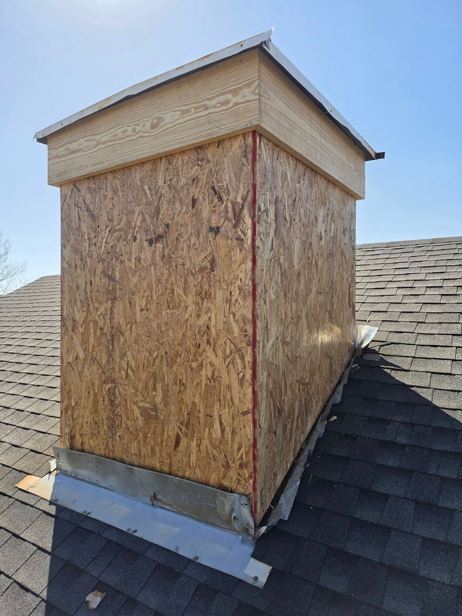 Rooftop chimney chase wrapped in OSB, with metal flashing and asphalt shingles around it.