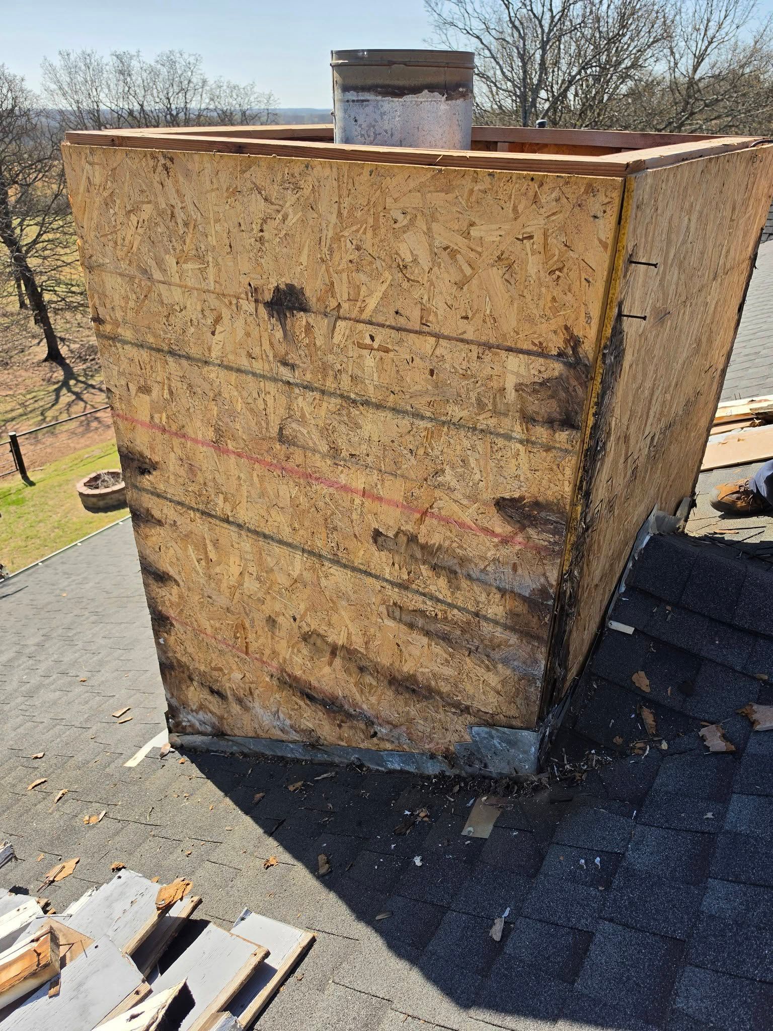 Damaged chimney stack on a shingled roof, with exposed plywood siding and a metal vent pipe.