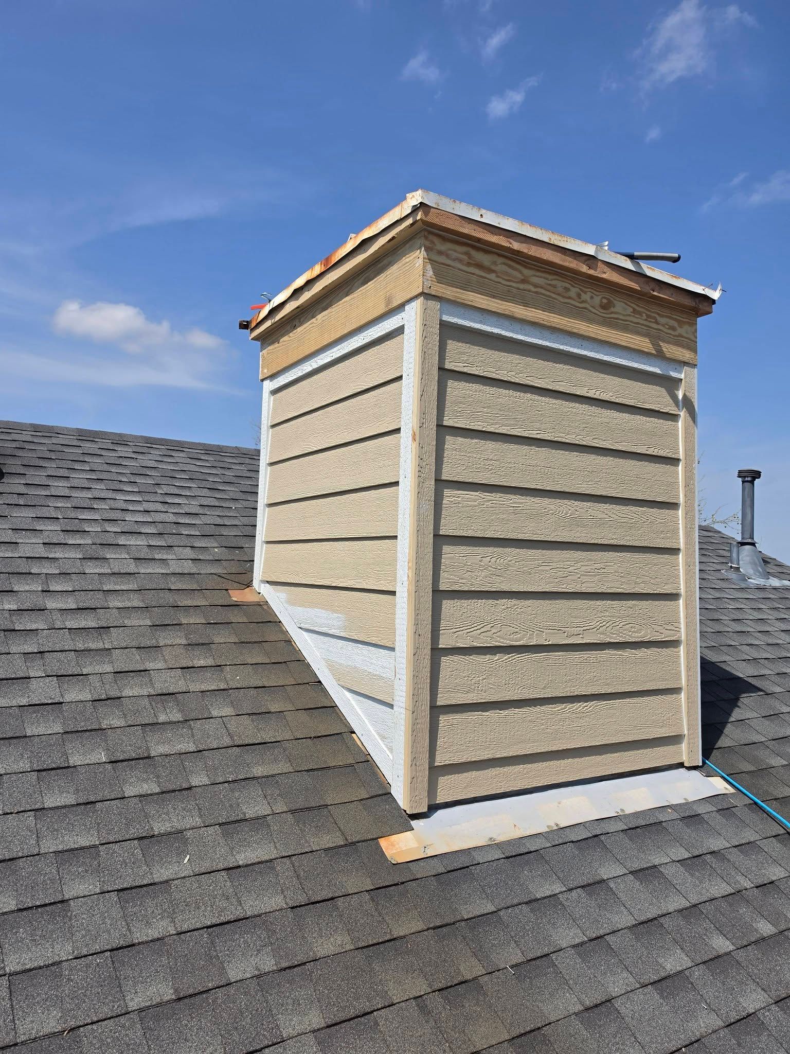 Rooftop chimney with beige siding on dark shingle roof under a blue sky.
