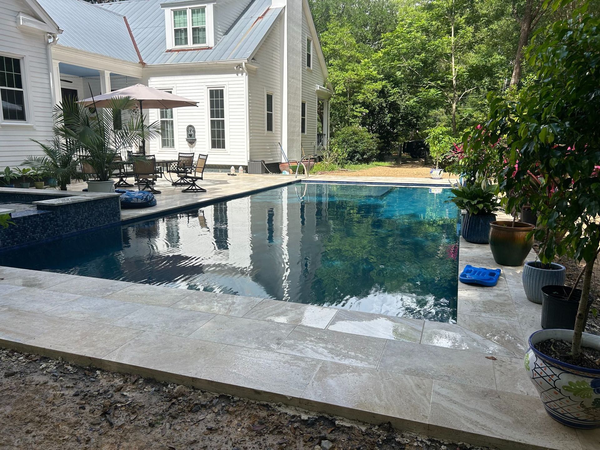 Backyard pool beside a house, with patio furniture and potted plants in bright daylight