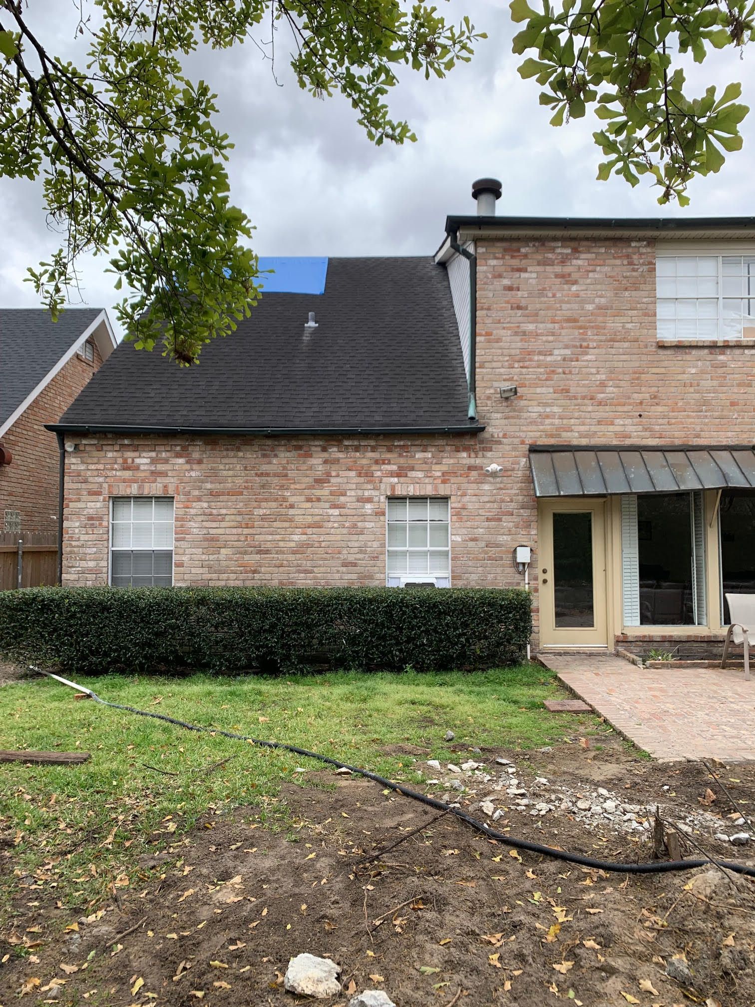 Brick townhouse exterior with dark roof, front hedge, and muddy yard under overcast sky