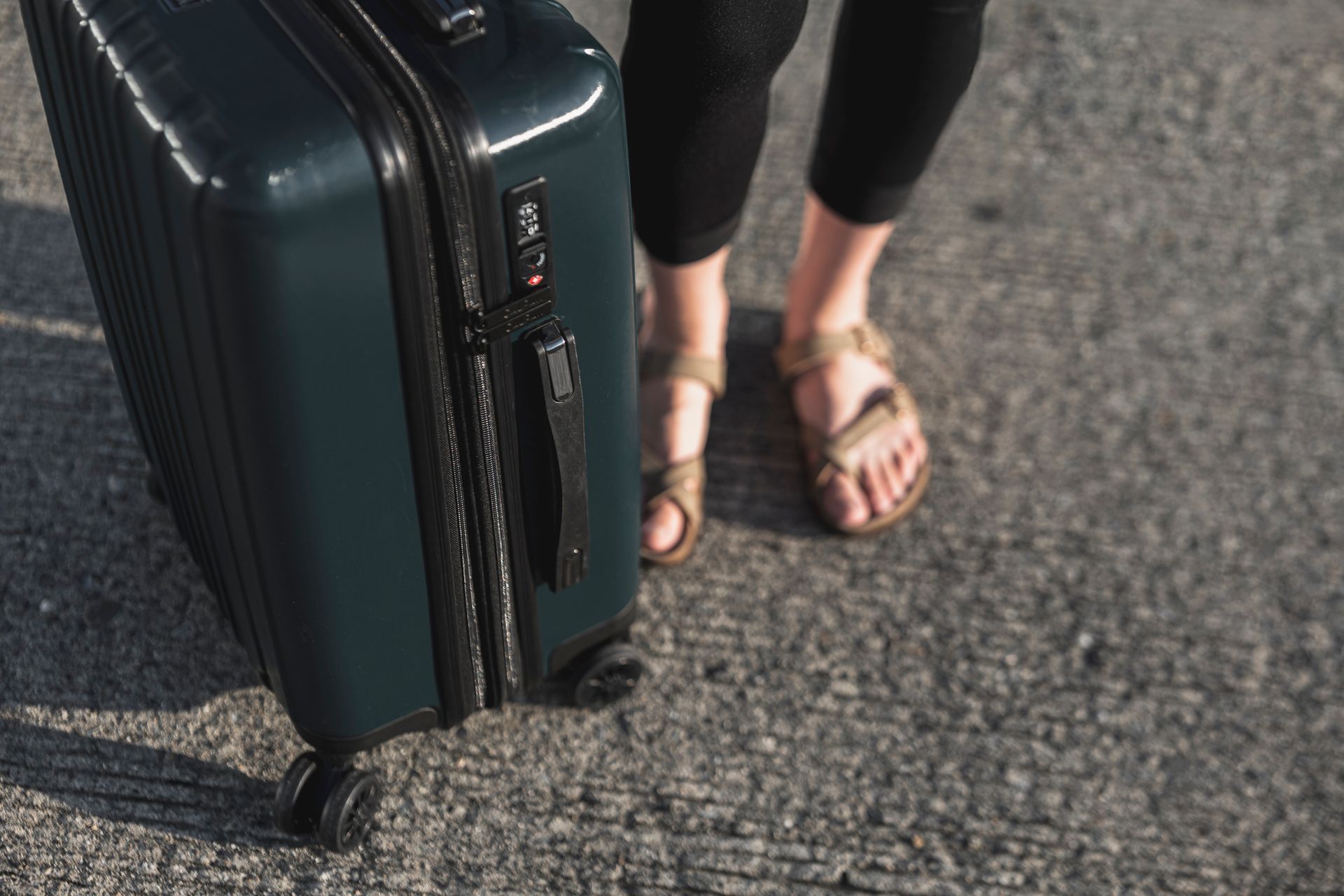 Person standing on concrete with a teal rolling suitcase; wearing sandals and black leggings.