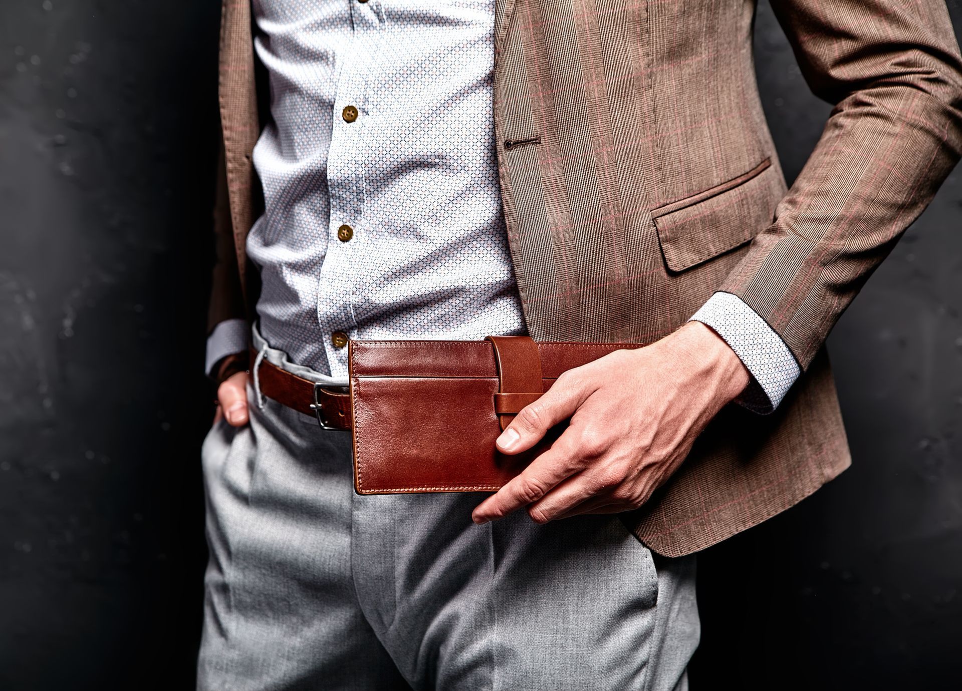 Man in suit holding a brown leather wallet on his hip. Dark brown jacket, light gray pants, and white patterned shirt.
