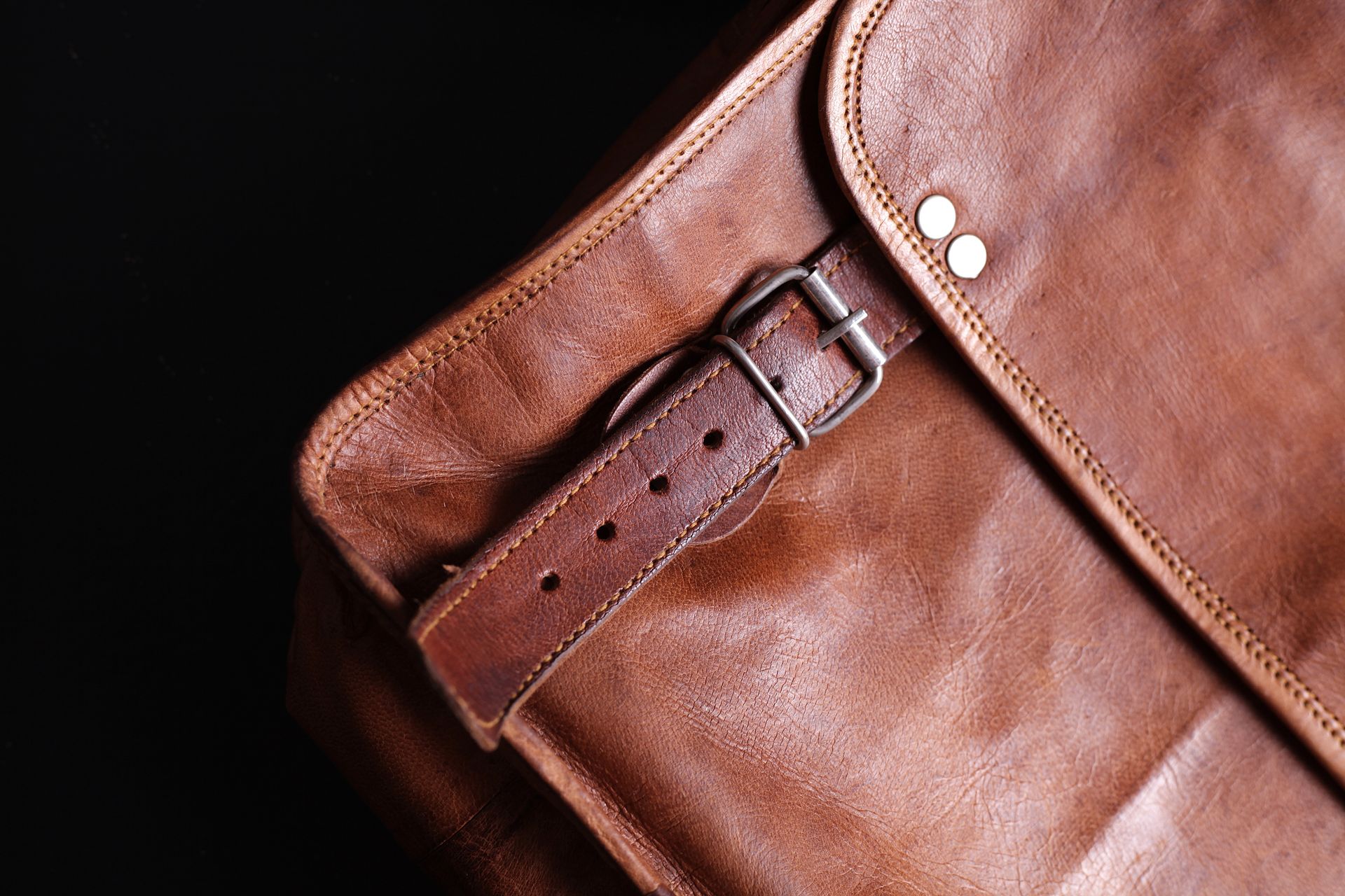 Brown leather satchel with buckle closure, rivets, and stitching, against a dark background.