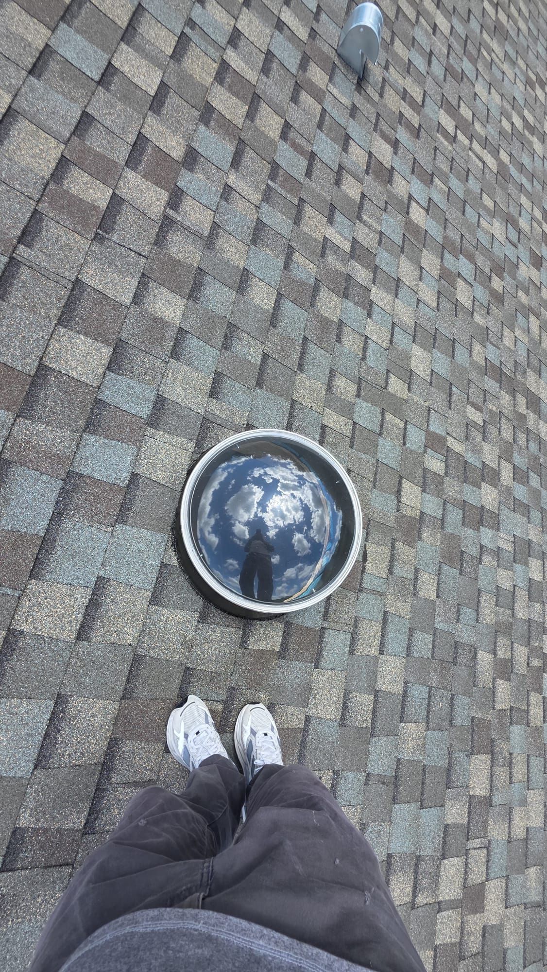 Looking down at white sneakers on a patterned shingle roof beside a round skylight.
