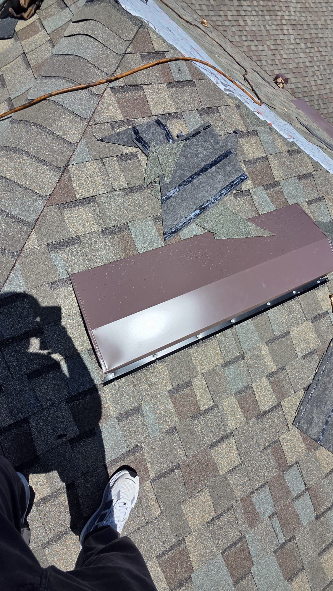 Person standing on a shingled roof beside a long brown metal panel and cables