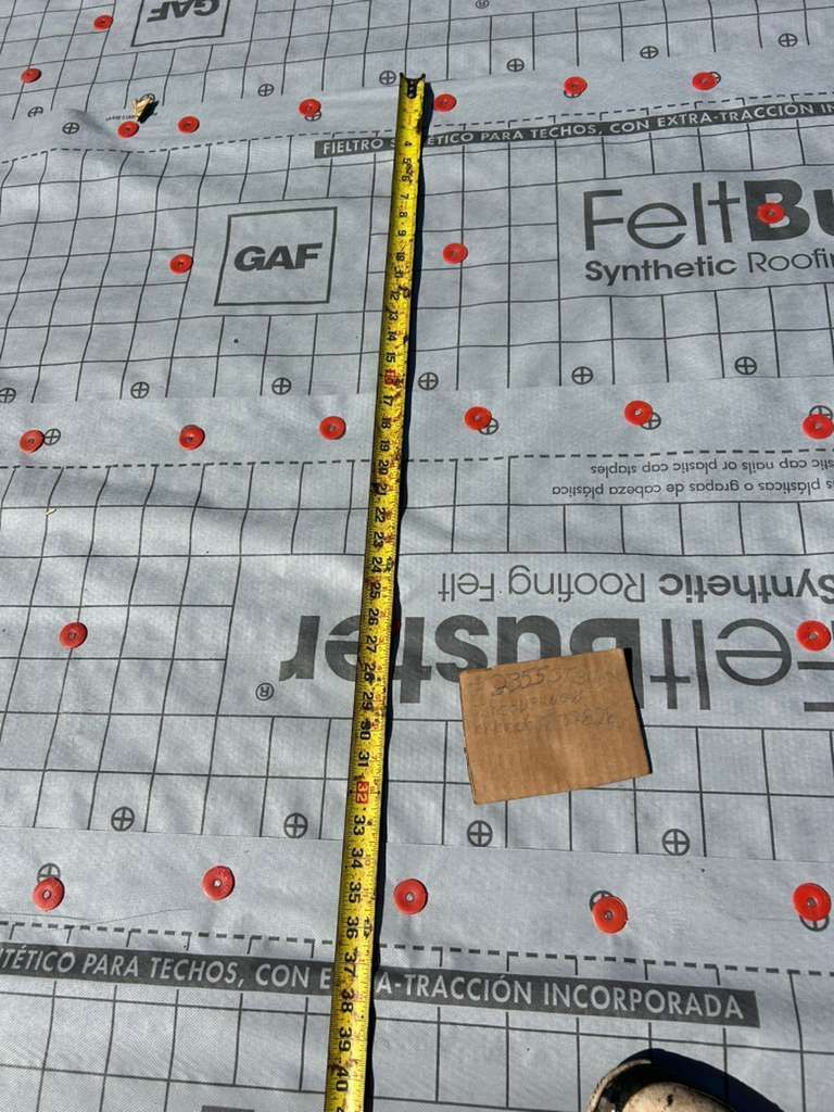 Tape measure on gray roofing underlayment with red markers and a small brown sample square.