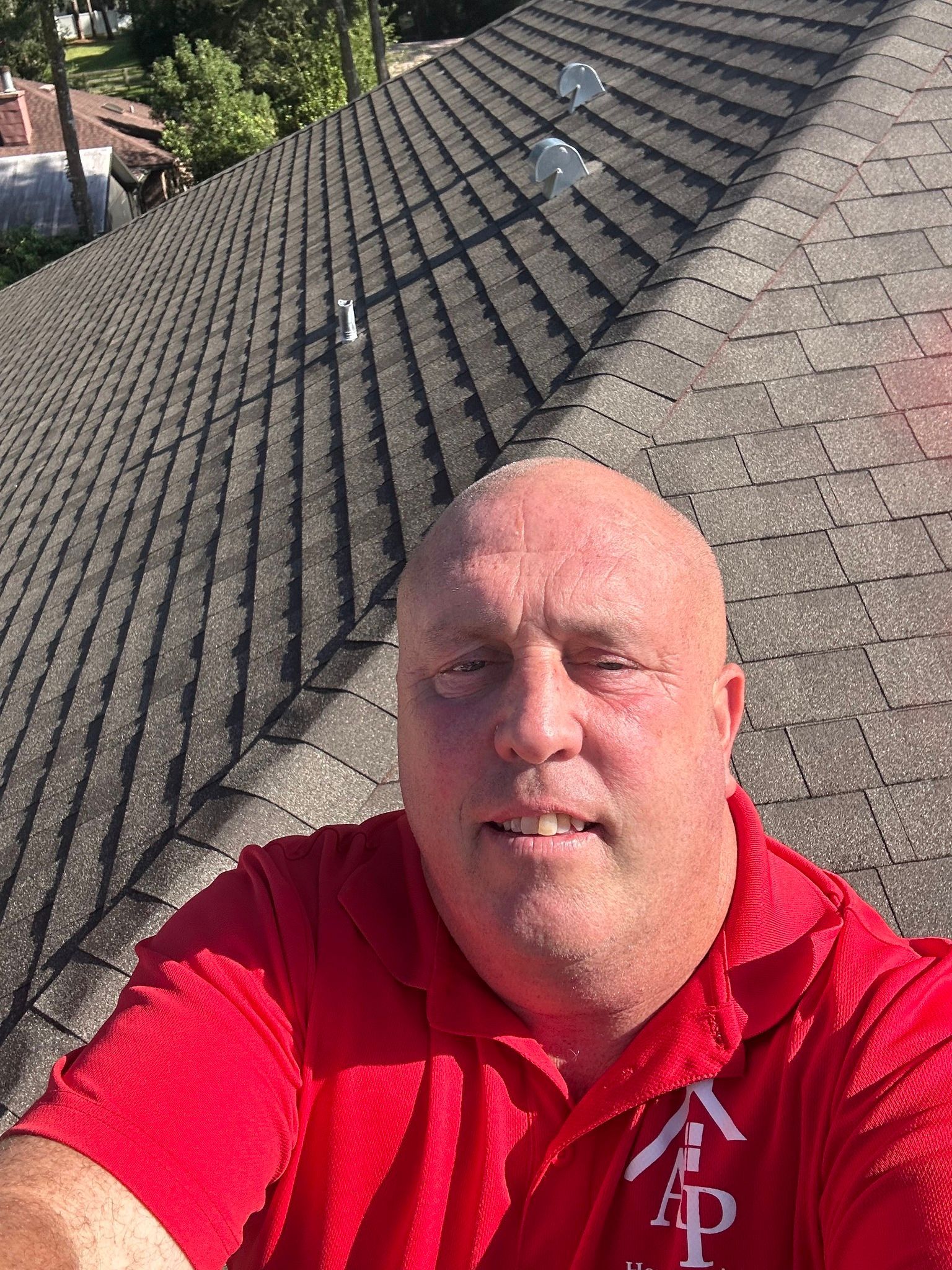 Man in a red shirt taking a selfie on a rooftop with gray shingles and trees in the background