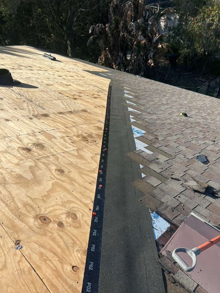 Roof under construction with plywood sheathing and shingles, showing a roofing ridge and ladder edge