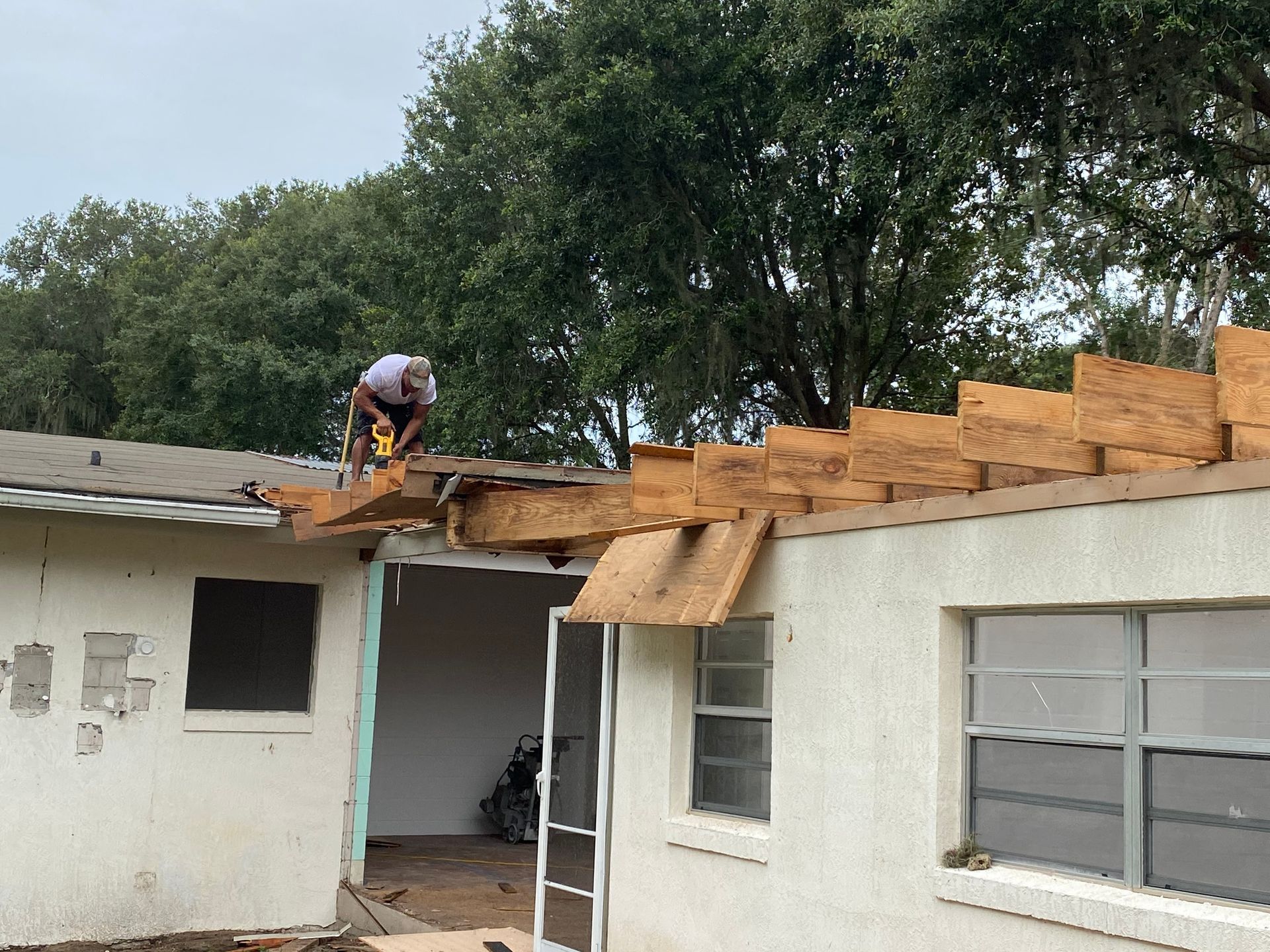 Worker repairing roof on a white house, with plywood exposed and trees behind it