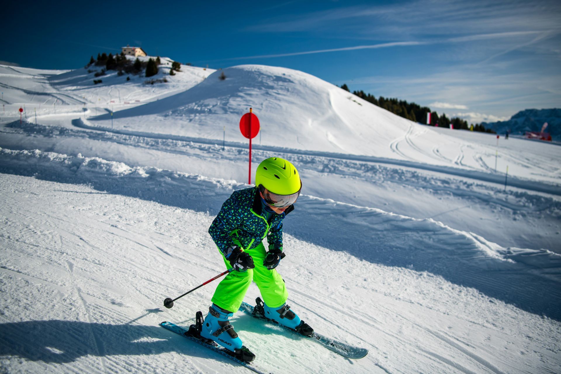 Child skiing downhill on a snowy slope, wearing a neon green helmet and ski outfit, with a red marker behind.
