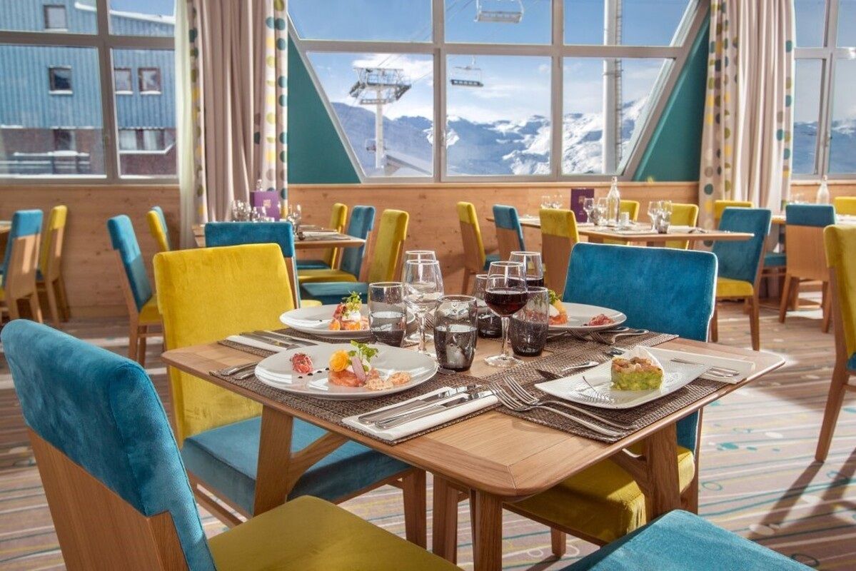 Colorful restaurant dining table with plated food, drinks, and blue-and-yellow chairs by large windows.