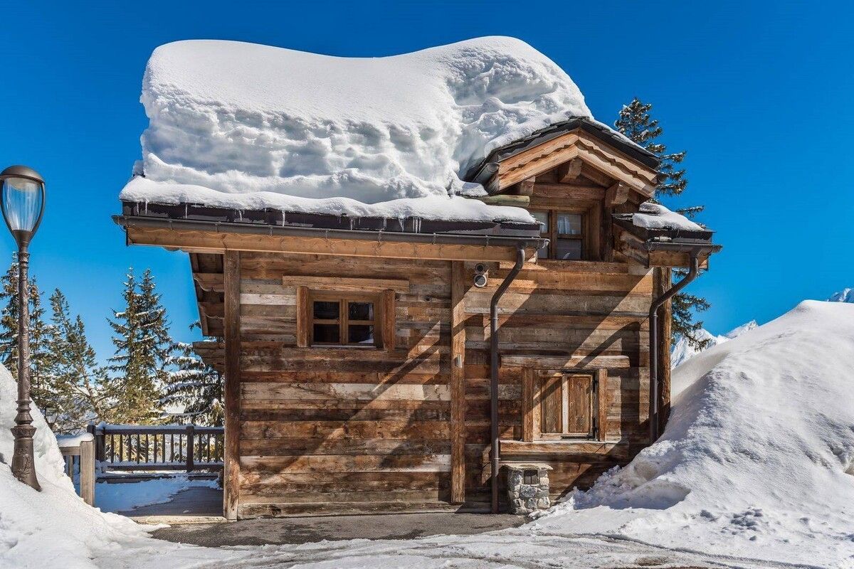 Snow-covered wooden cabin under a bright blue sky, with a large snowdrift beside it.