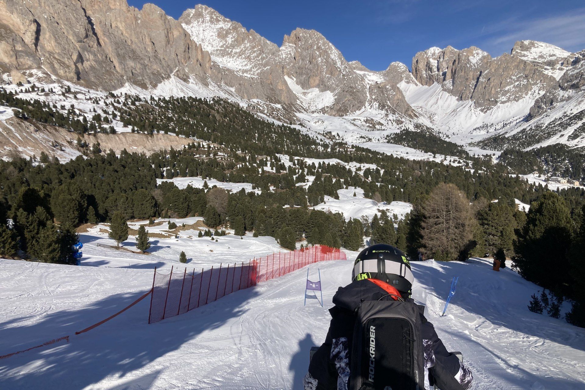 Skier descending a snowy mountain trail with pine trees and rugged peaks under a clear sky