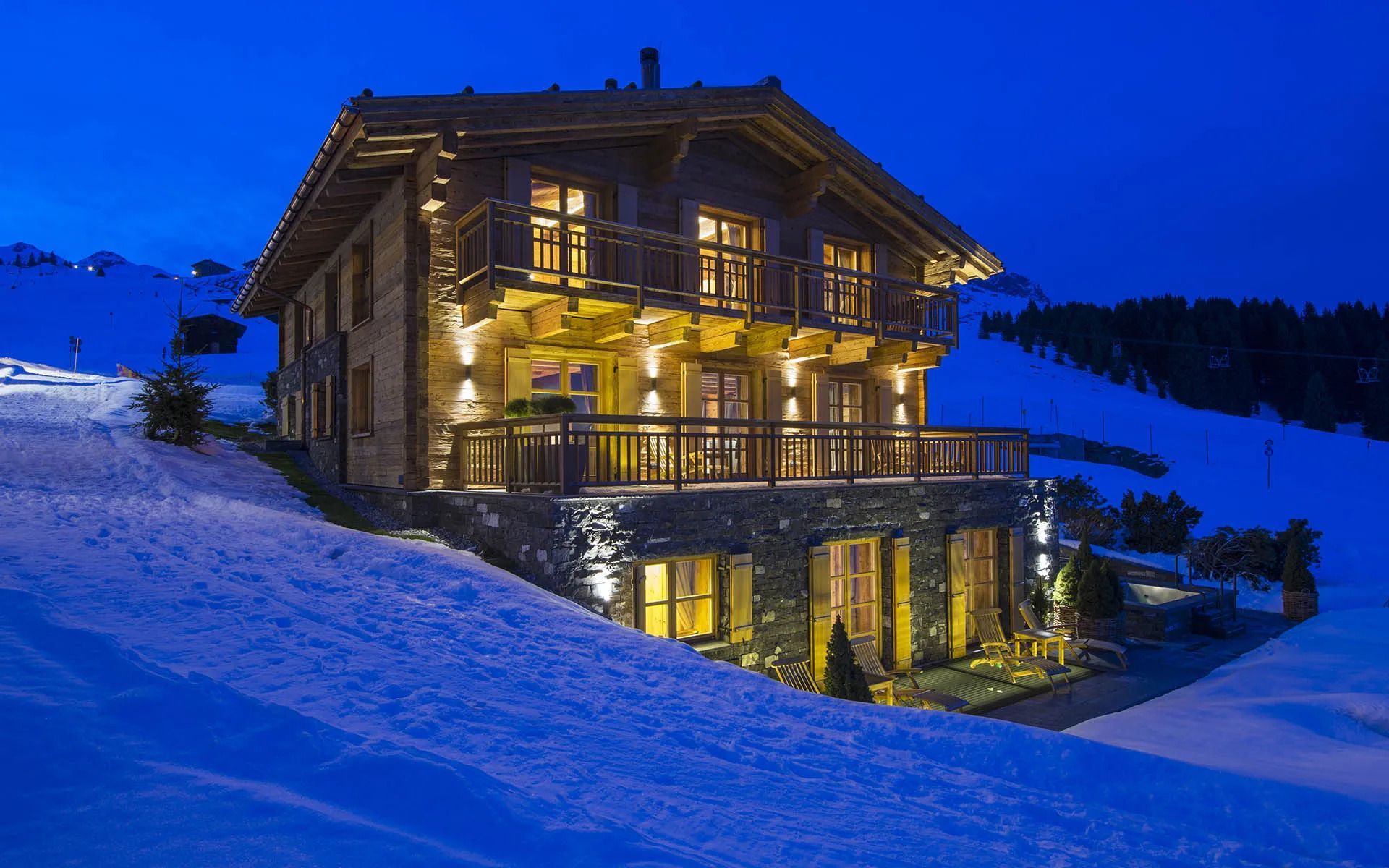 Wooden chalet glowing warmly on a snowy hillside at dusk, with lights reflecting in the windows