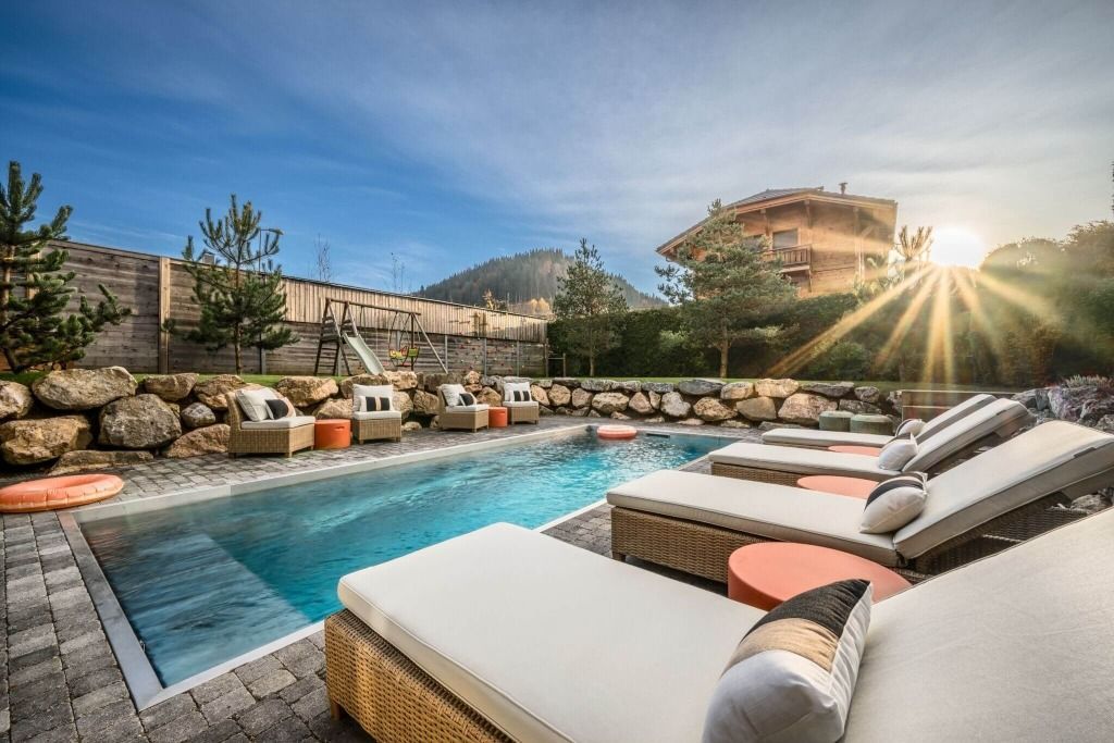 Modern poolside lounge with white chairs, turquoise water, stone deck, and sun flare at sunset