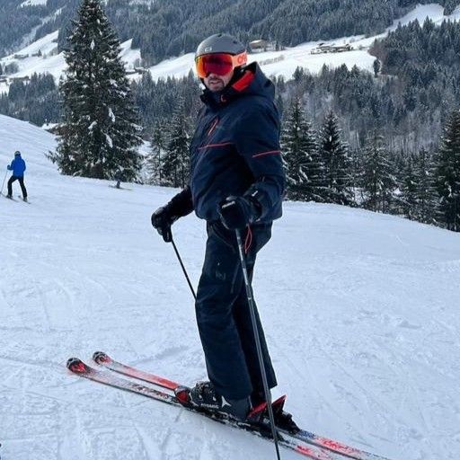 Skier in dark gear and red goggles standing on a snowy slope with trees and mountains behind.