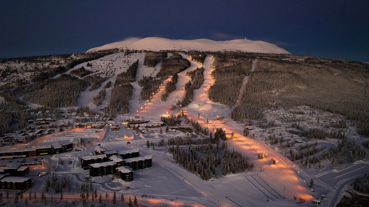 Snow-covered ski slope at dusk, with lit trails winding down a mountain above buildings and trees.