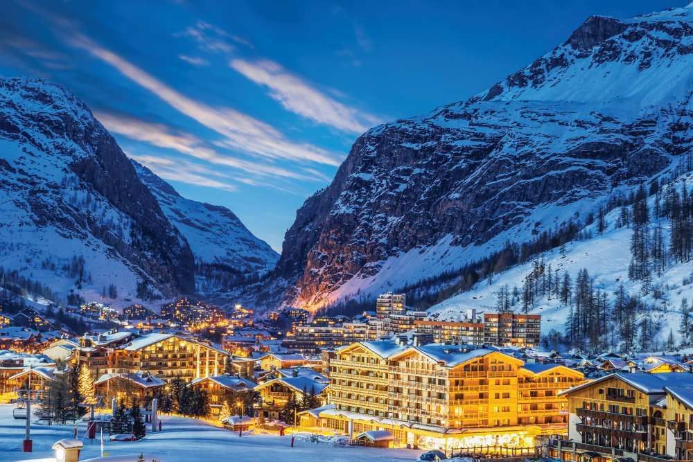 Snowy alpine village at dusk, warmly lit buildings nestled between steep mountain slopes under a blue sky