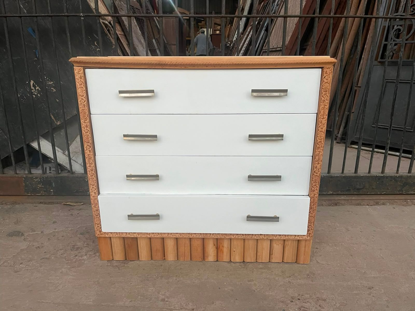 Cómoda blanca de cuatro cajones con detalles y tiradores de madera, fotografiada al aire libre frente a una valla metálica negra.