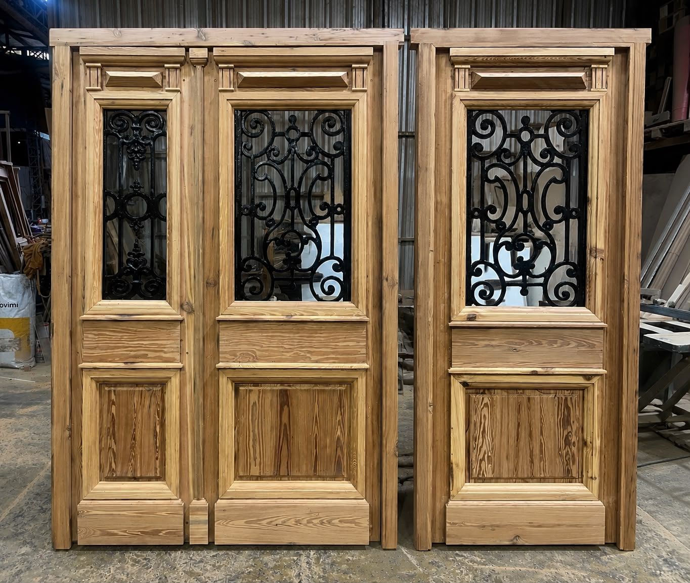Tres puertas de madera con paneles de vidrio de hierro forjado negro ornamentados, expuestas al aire libre en un taller.