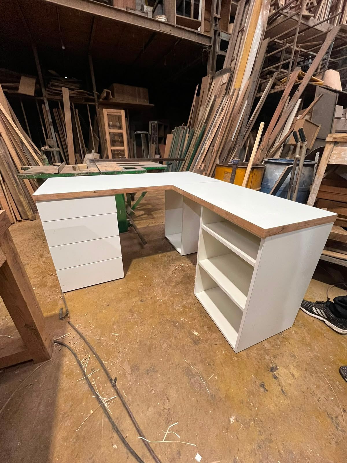 Escritorio blanco en forma de L con cajones y estantes en un taller de carpintería, rodeado de madera y herramientas.