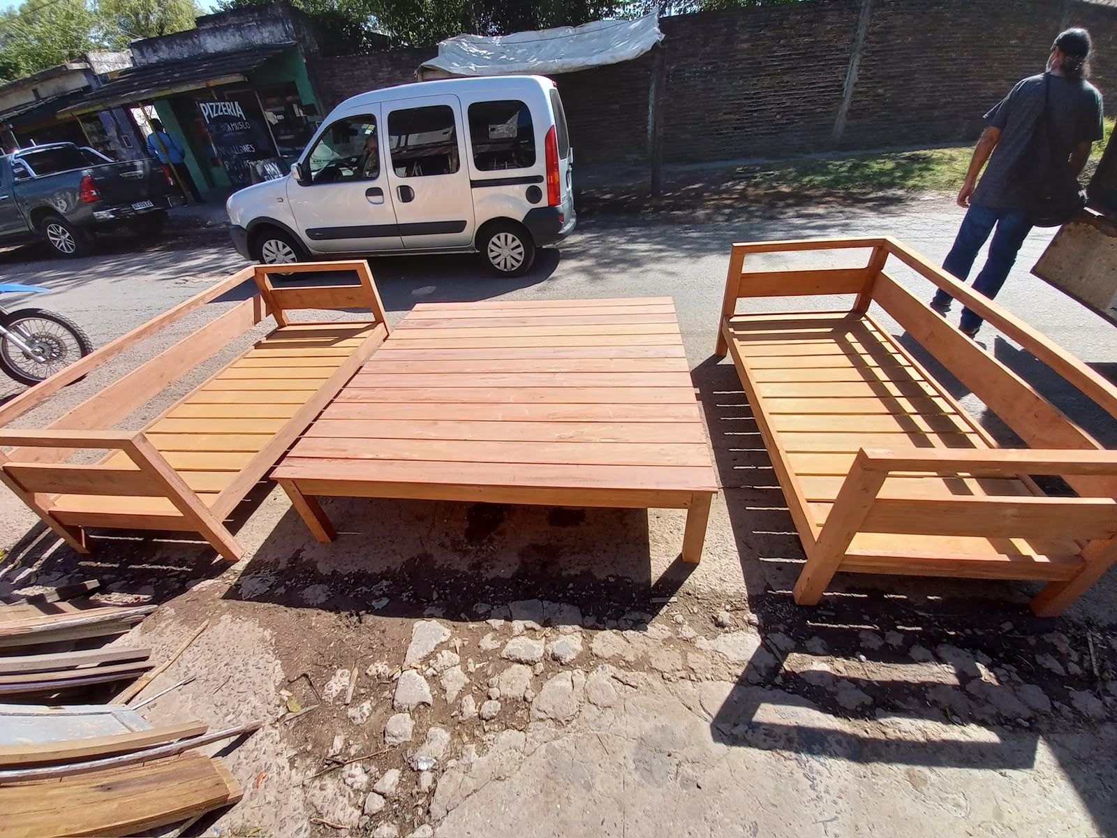 Conjunto de muebles de exterior de madera con dos bancos alrededor de una mesa baja, aparcado cerca de una furgoneta blanca en una calle.