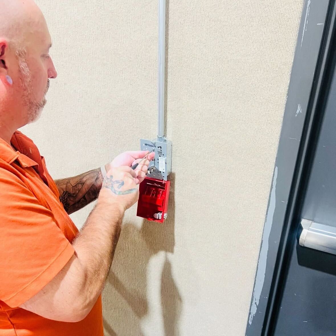 Man in orange shirt attaching a red fire alarm device to a wall panel