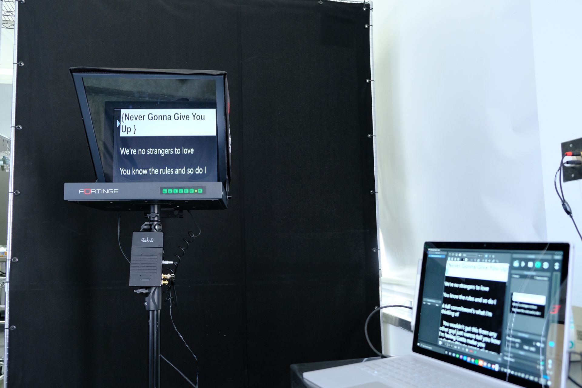 Teleprompter setup with black backdrop, monitor displaying text, and laptop showing editing software.