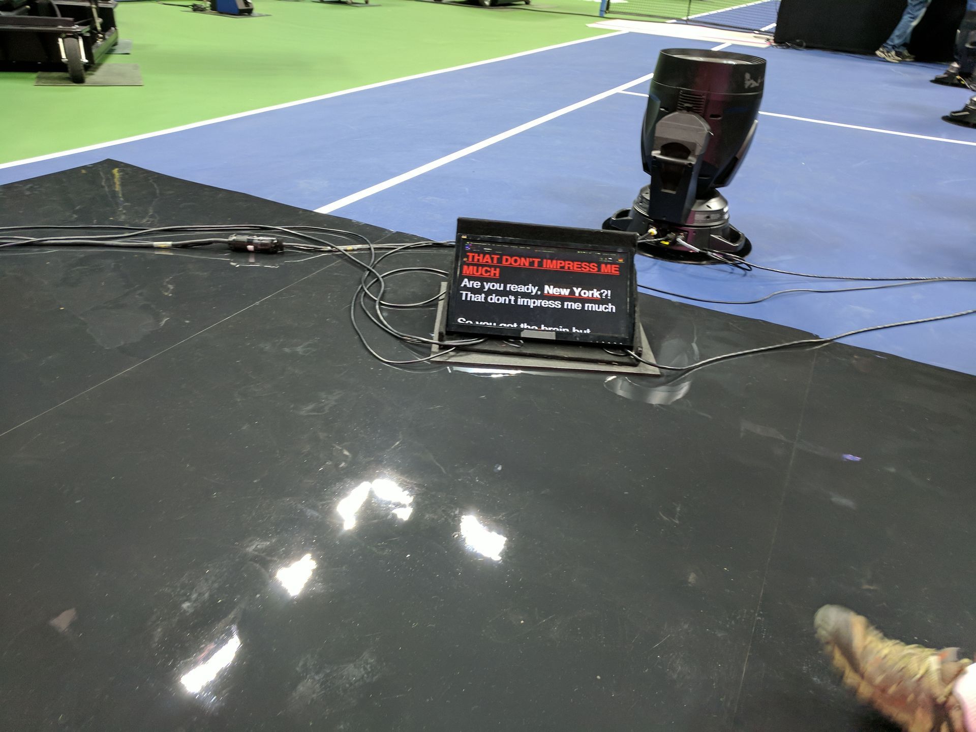Stage floor with monitor displaying text, cables, spotlight, and tennis court in background. Teleprompters