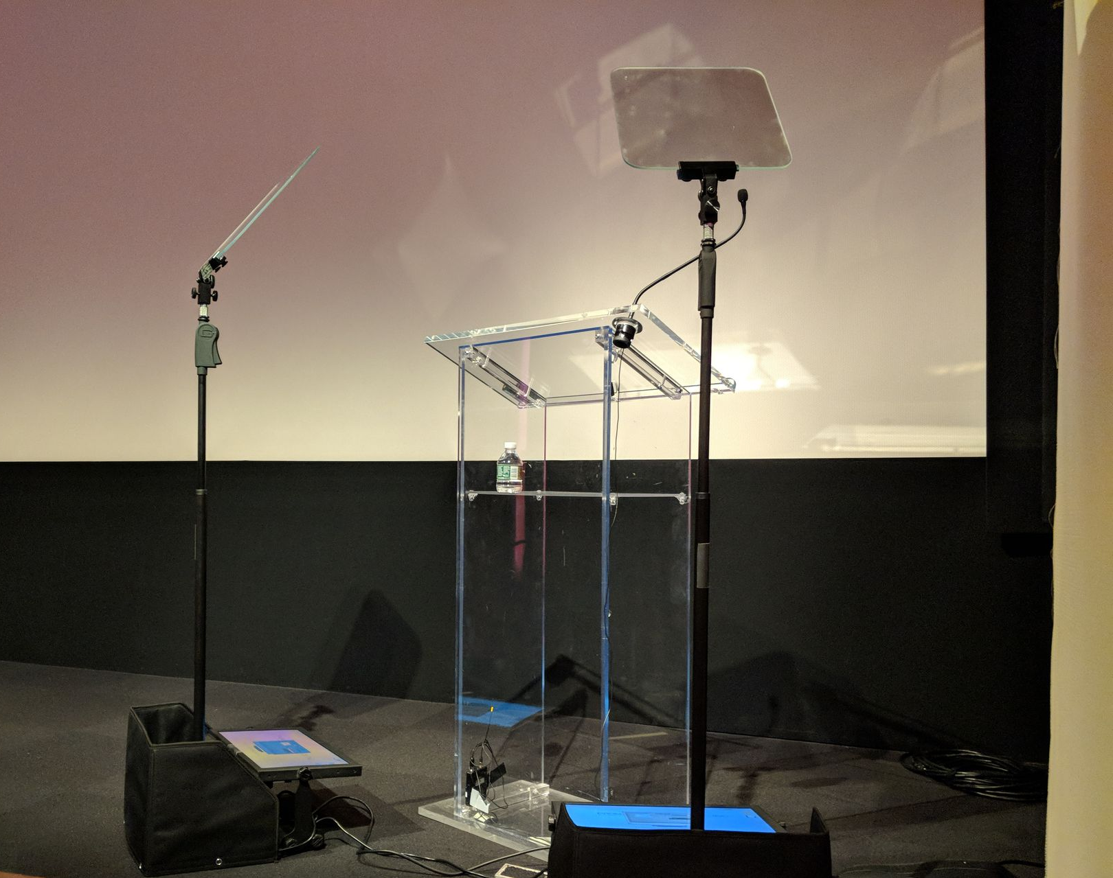 A clear lectern with a transparent top and two microphones set up in front of a screen. Teleprompters Shine