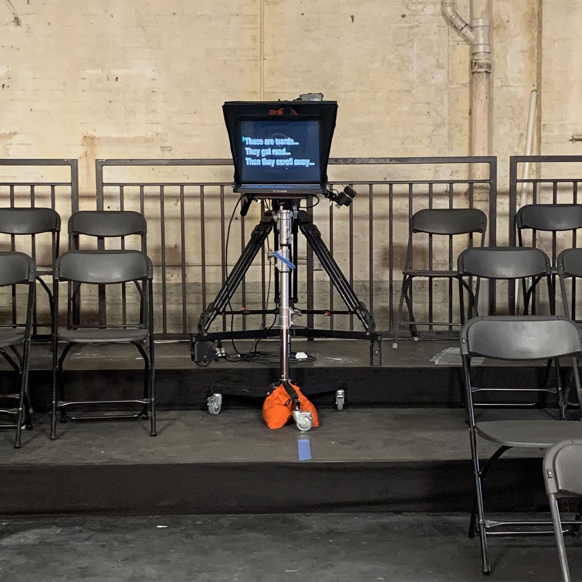 Teleprompter on tripod; rows of empty chairs on a raised platform against a wall.