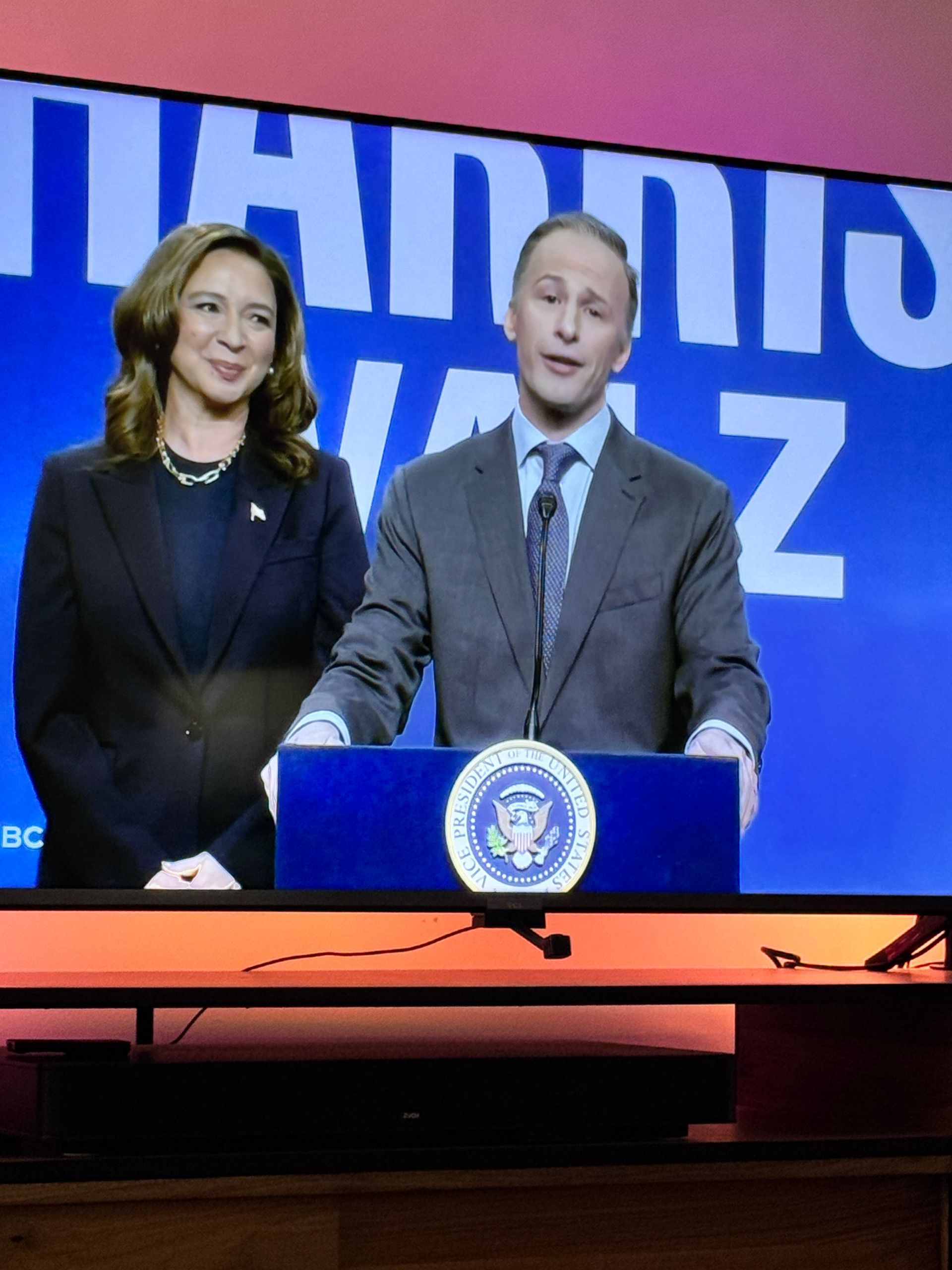 Two SNL People stand behind a podium. A blue sign reads 