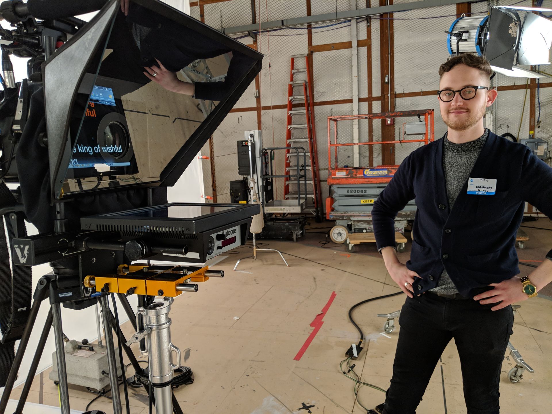 Man in a studio with a teleprompter and camera. He smiles, hands on hips, wearing a blue cardigan.