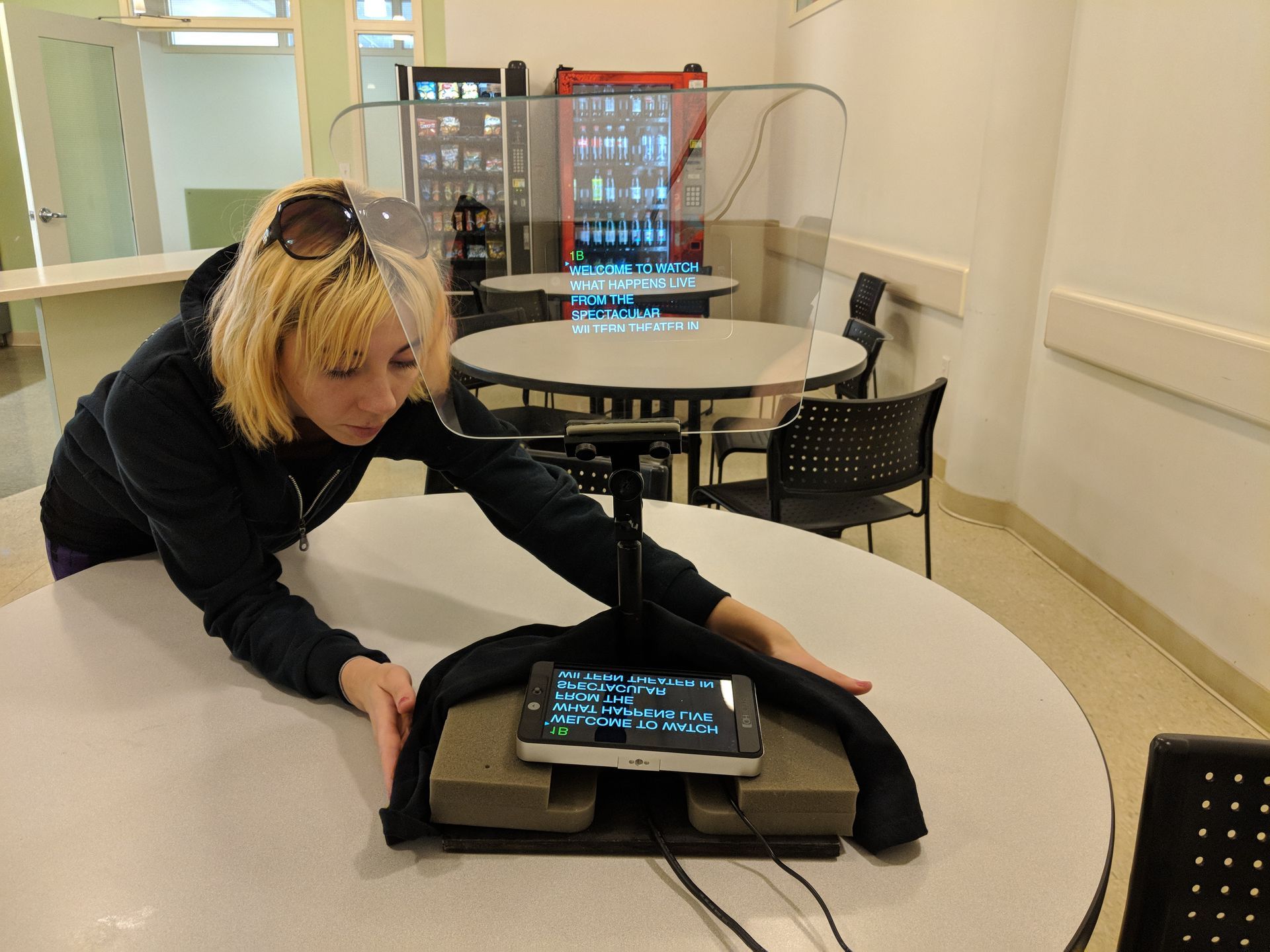 Woman interacting with a phone teleprompter on a table. Tables, chairs, and a vending machine are in the background.