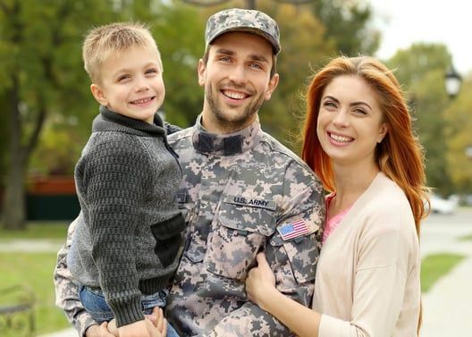 Soldier in uniform holds his son; wife smiles beside them, outdoors.