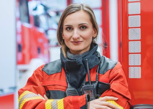 Woman firefighter in red uniform stands with arms crossed in front of a fire truck.