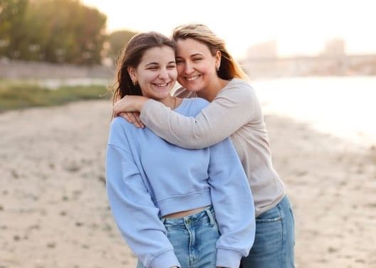 Mother and daughter embrace on a beach, smiling. Sunlight bathes them.