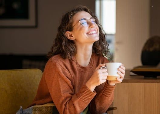 Woman with curly hair, glasses, smiles holding coffee mug, enjoys sunlit space.