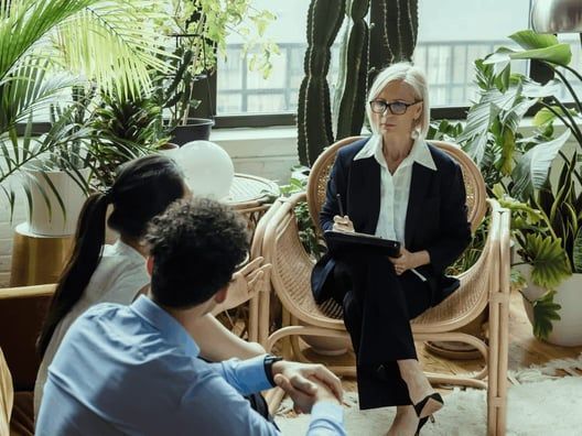 Therapist in a blazer takes notes, facing a couple seated in front of her. Room is filled with plants.