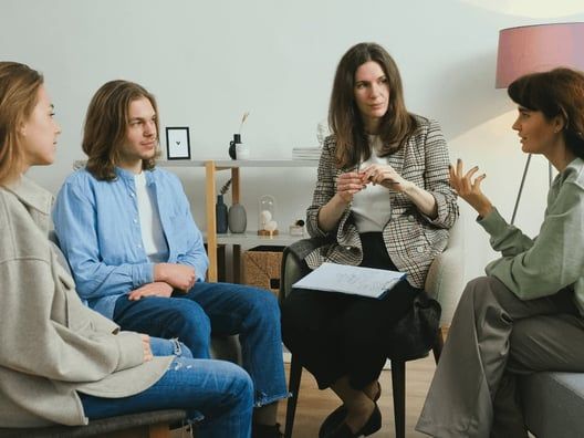 Four people in a counseling session; a counselor in a blazer talks while the other three listen in a circle.