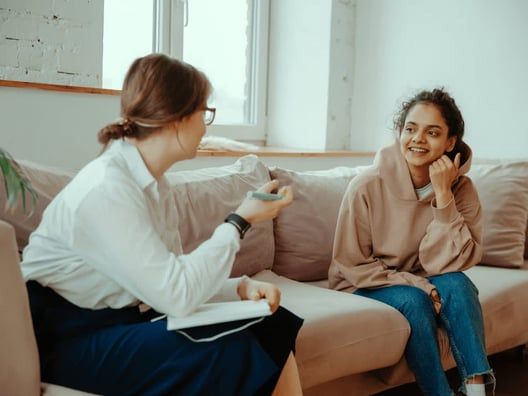 A therapist in a white coat gestures while speaking to a smiling young woman in a beige hoodie on a couch.
