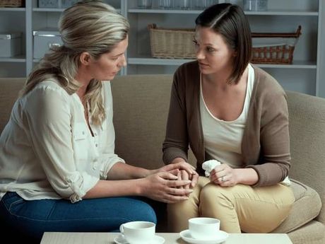 Two women sitting on a couch, one holding the other's hands, looking concerned.  White cups sit on the table in front of them.