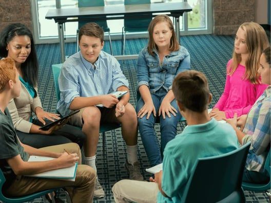 A group of teens in a circle, seated in chairs, engaged in a discussion, possibly a group therapy setting.
