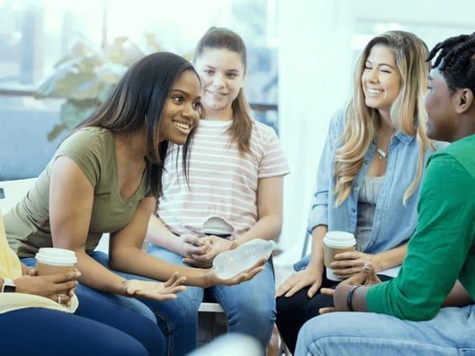 A diverse group of women sitting in a circle, smiling and talking, holding coffee cups. They appear to be in a supportive group setting.
